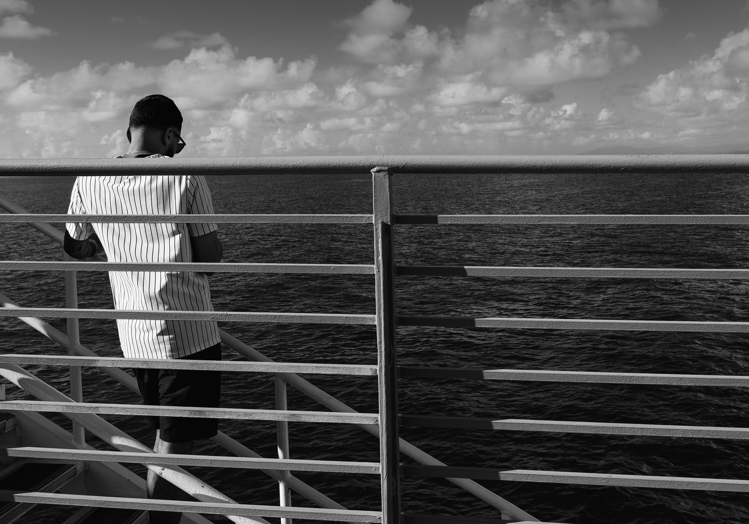 Black and white rear view of a man in striped shirt standing at a ship railing overlooking the ocean.