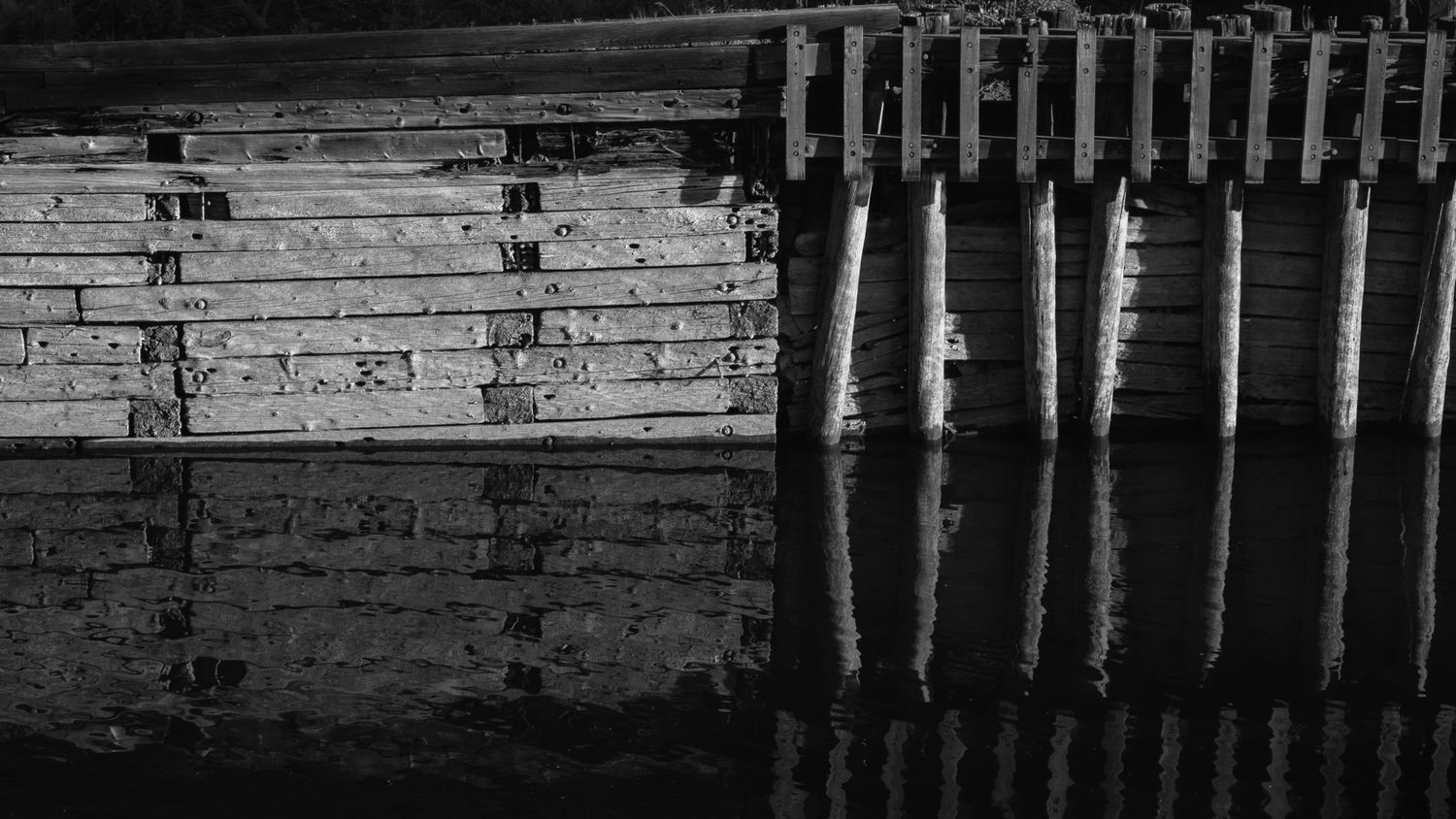 Black and white close-up of weathered wooden dock planks and pilings reflected in dark still water.