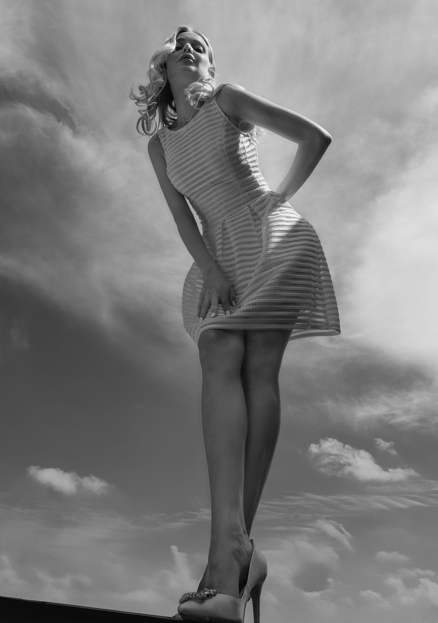 Black and white low-angle shot of a blonde woman in striped dress and heels against dramatic sky.