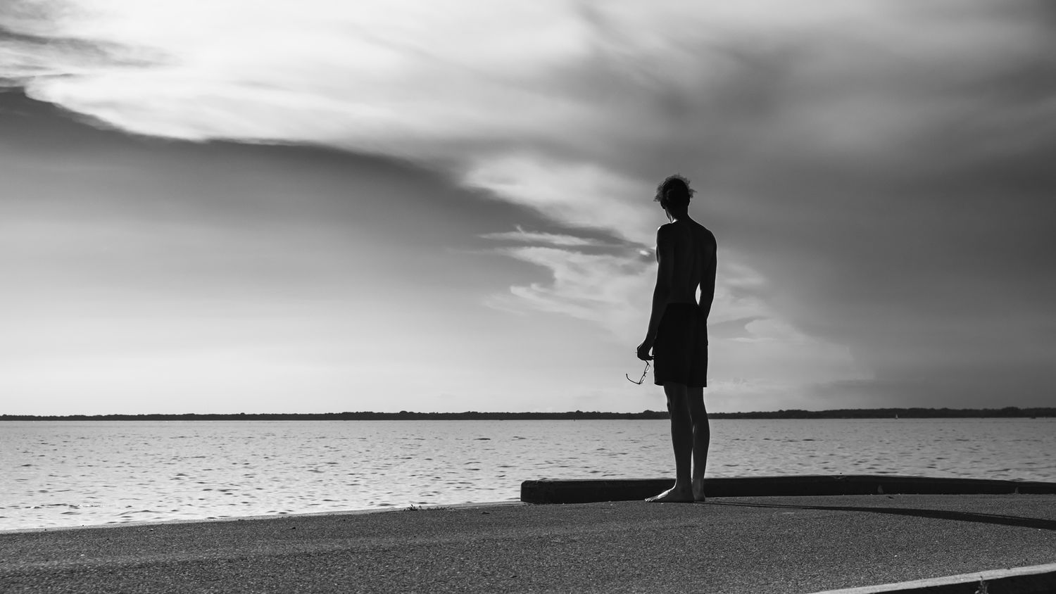 Black and white photo of a lone person standing on a waterfront promenade gazing at the sea under dramatic cloudy skies.