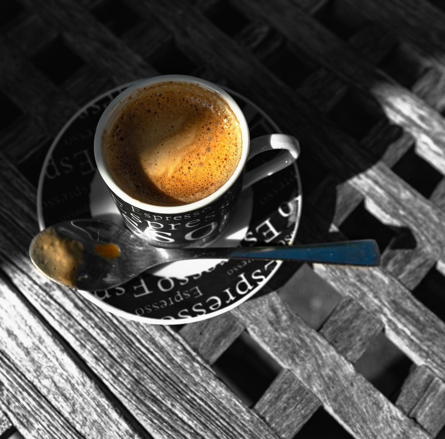 Selective color photo of an espresso coffee cup on a saucer atop a wooden slatted table.