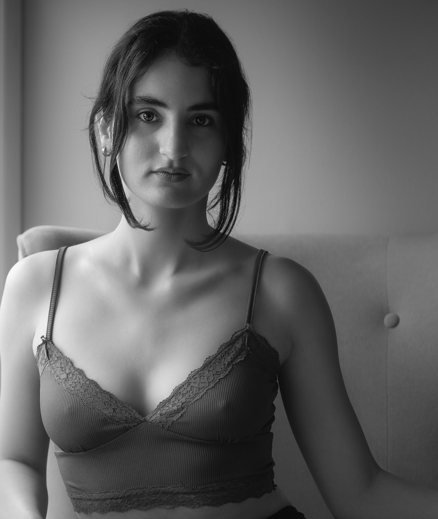 Black and white portrait of a young woman in lace bralette seated on a light-colored chair.