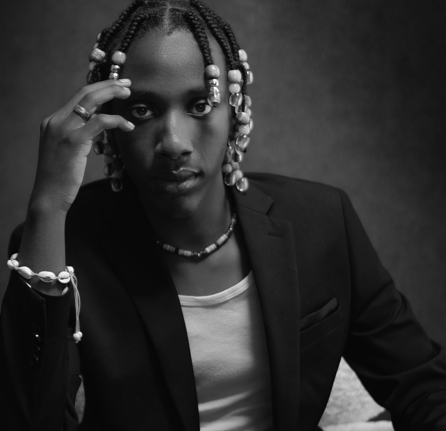 Black and white portrait of a young man with braided hair and beads wearing a blazer and necklace.