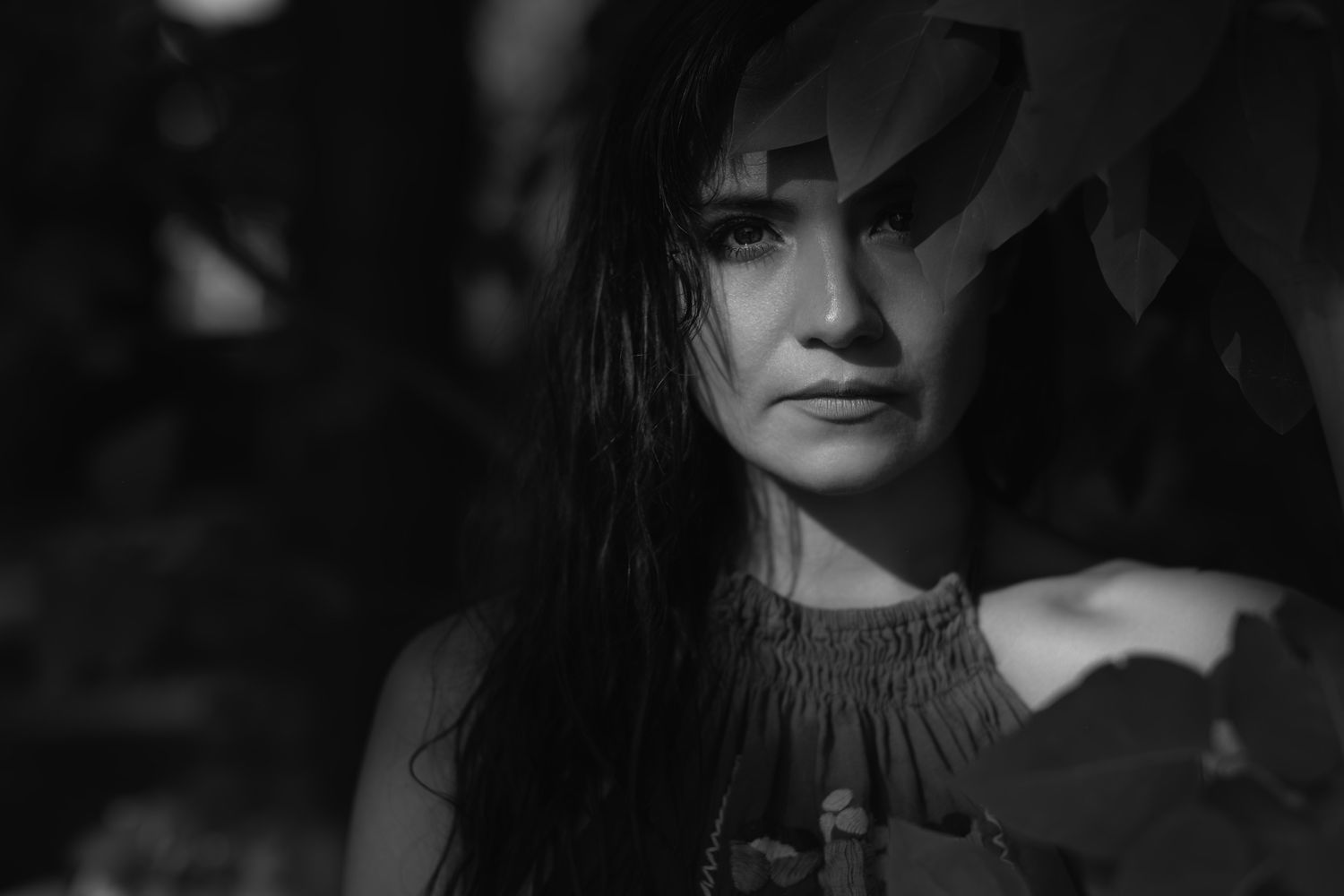 Black and white portrait of a young woman with long dark hair surrounded by leaves in soft natural light.