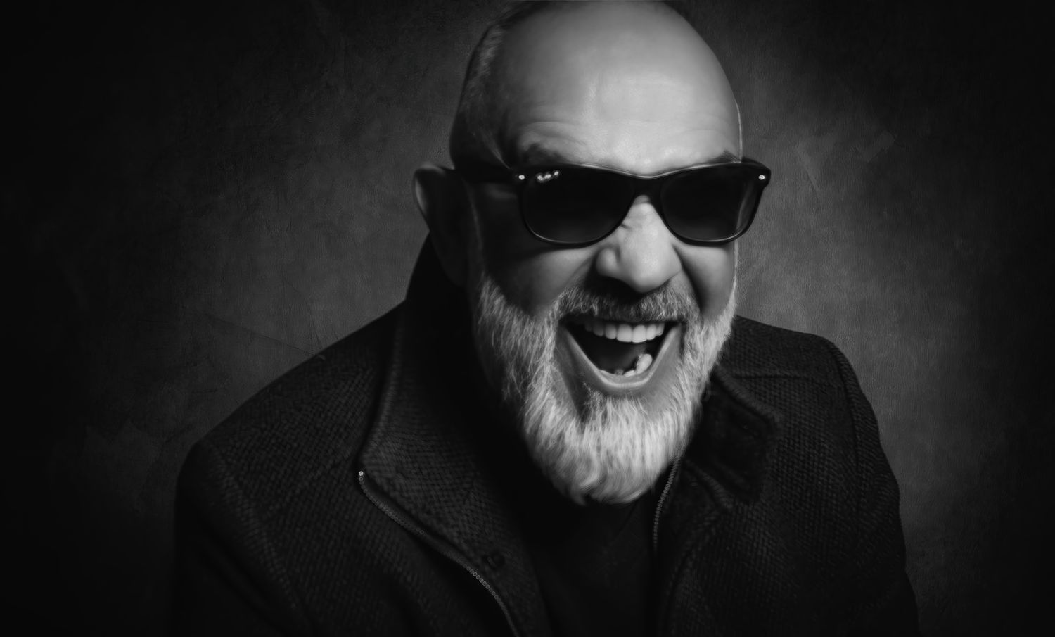 Bald man with white beard and sunglasses laughing in a black and white portrait photo.
