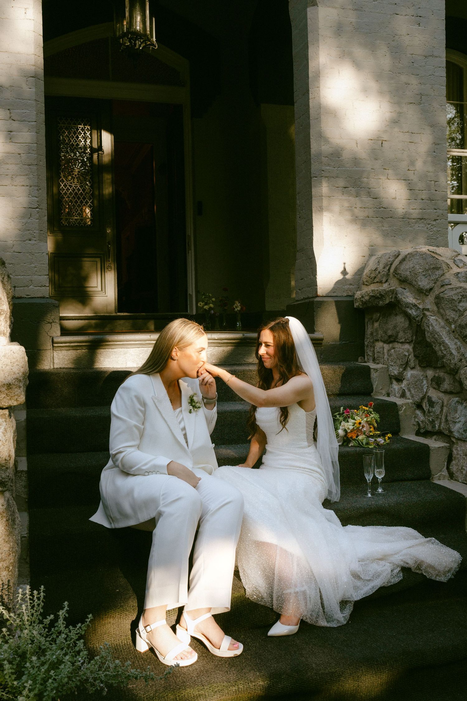 Two brides in white sitting on stone steps, sharing a tender moment in dappled sunlight.