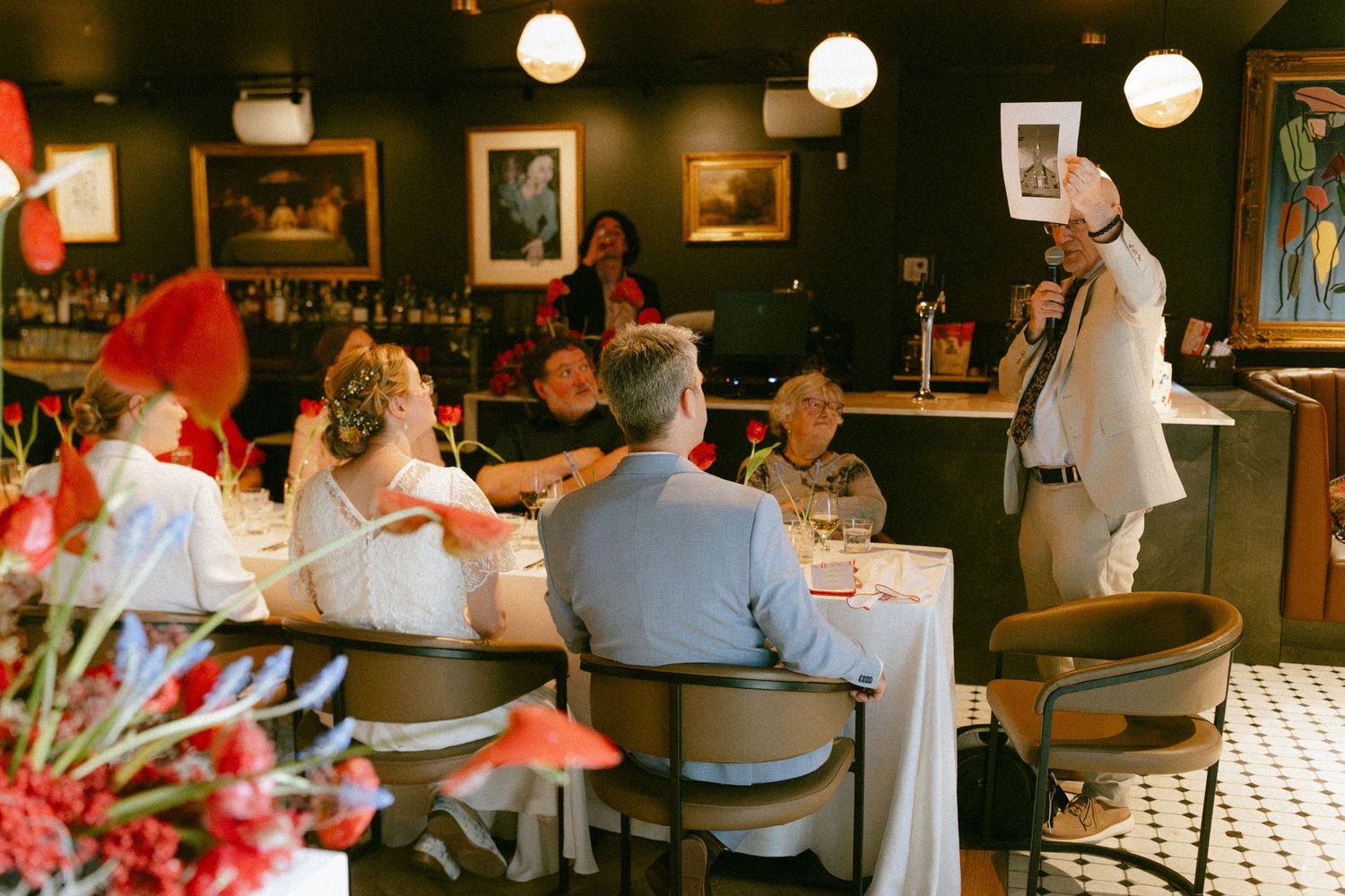 Wedding reception speech at elegant dark-walled restaurant with red floral decorations.