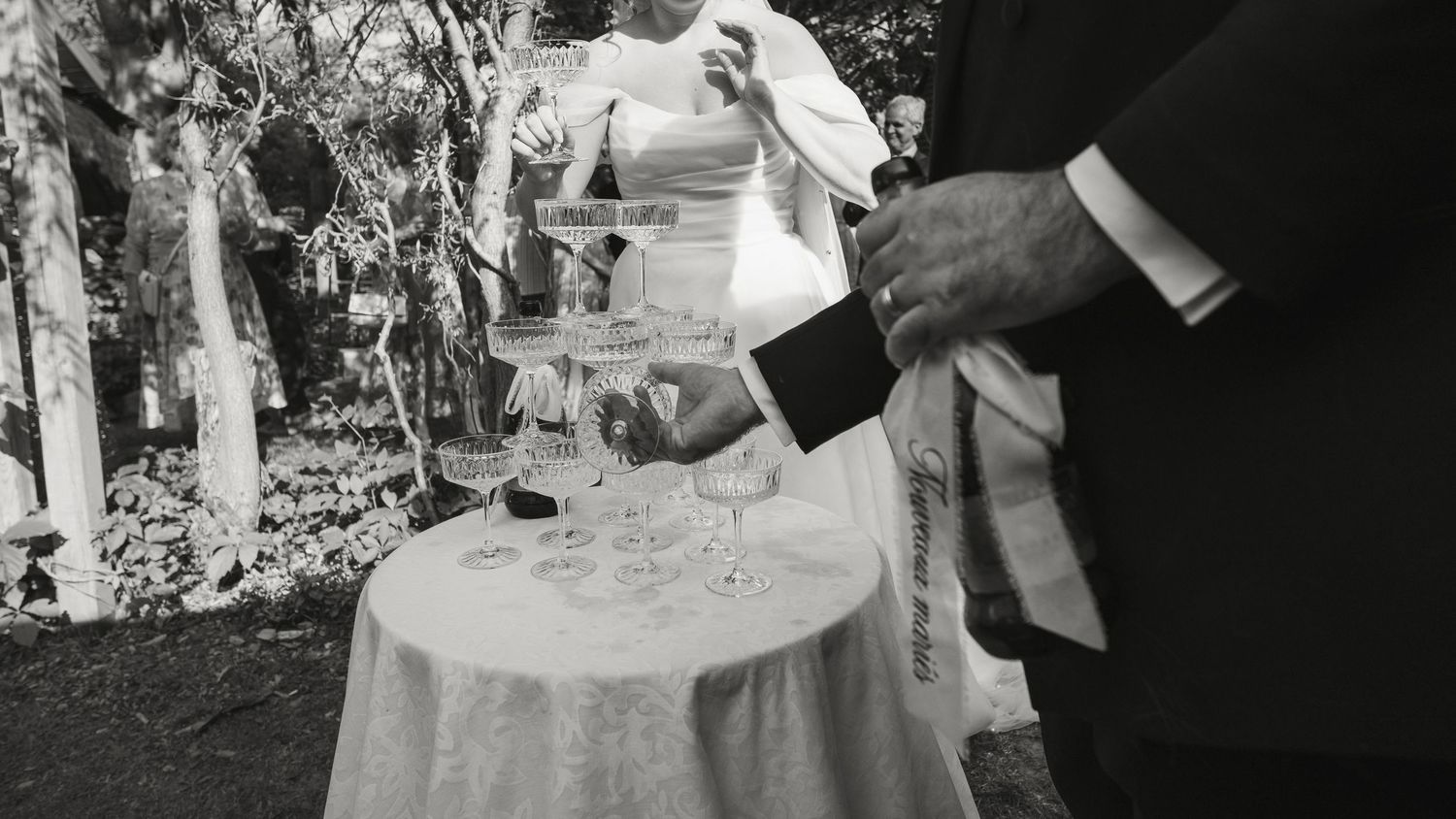 Black and white photo of champagne tower being poured at an outdoor wedding reception.