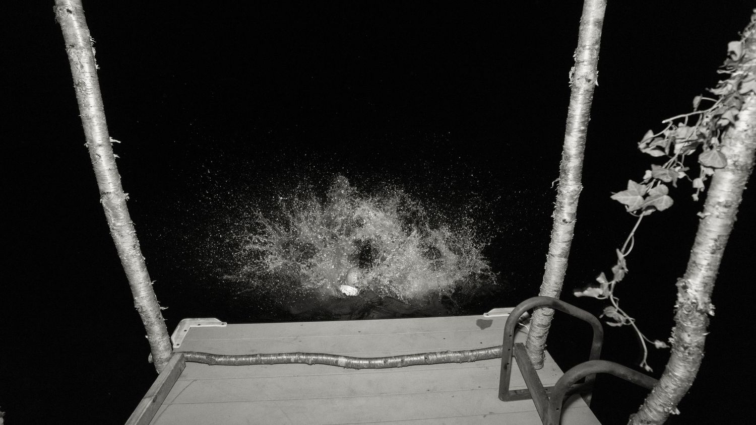 Black and white night photo of water splashing between two wooden posts on a deck with a tree nearby.
