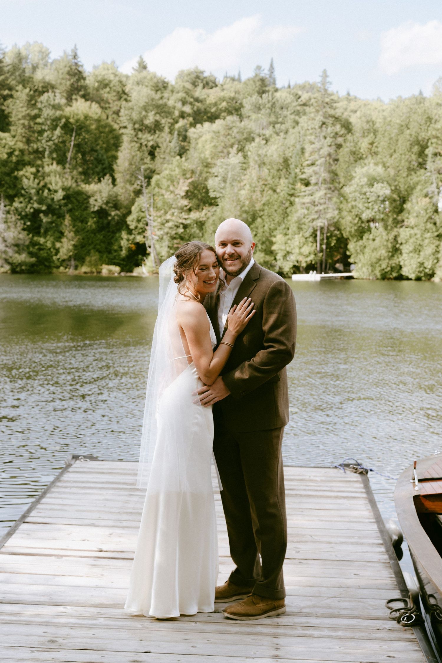 Bride and groom embrace on a wooden dock by a serene lake surrounded by lush green forest on their wedding day.