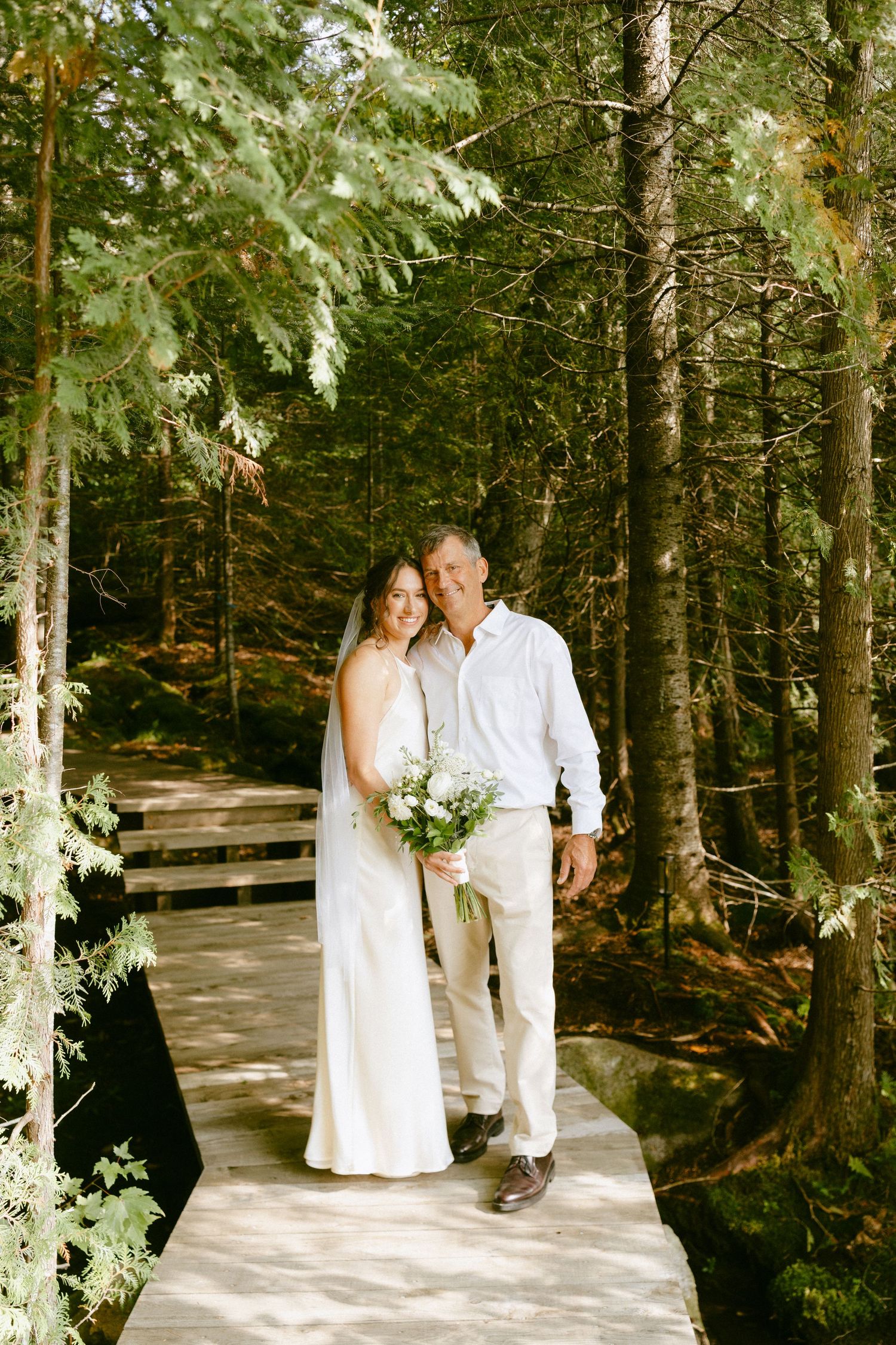 Bride and groom walking on woodland boardwalk, bride holding white floral bouquet.