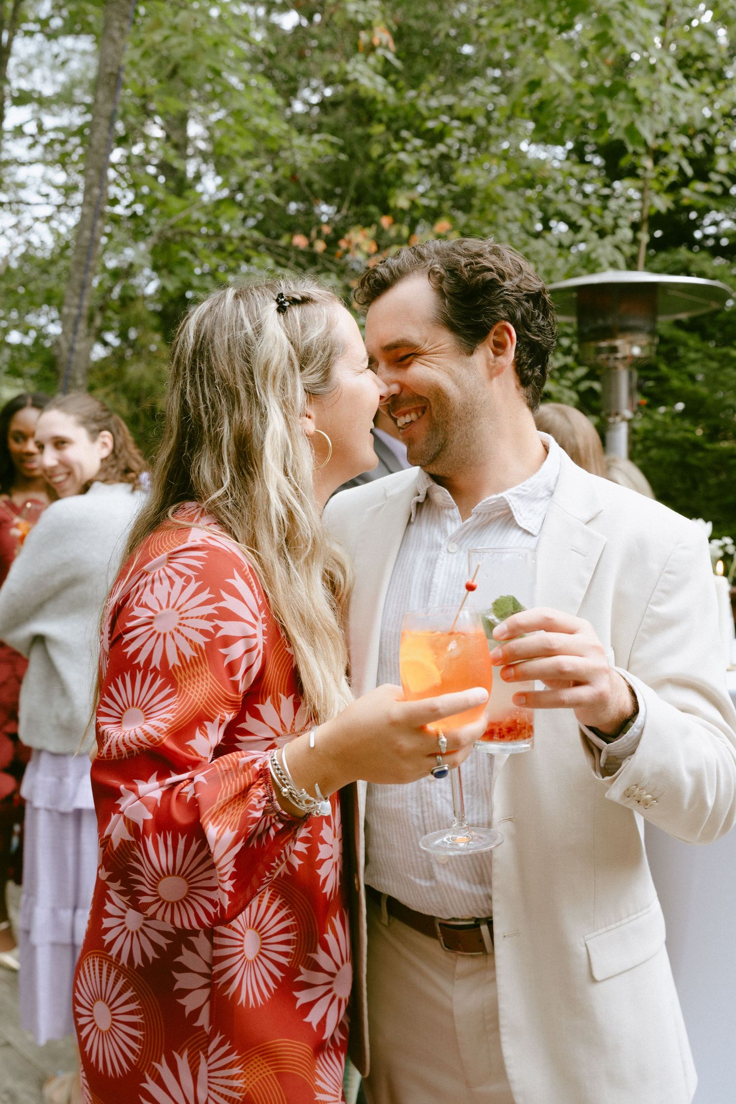 Smiling couple in festive attire toasting with Aperol spritz cocktails at outdoor wedding reception.