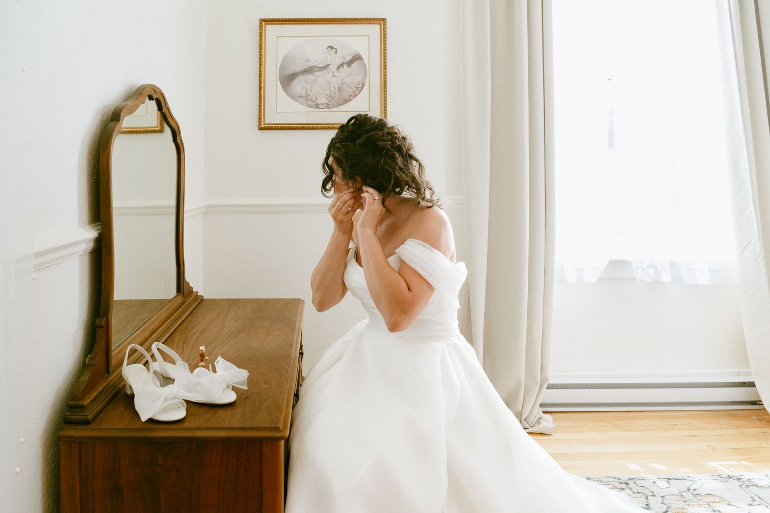 Bride in off-shoulder white gown adjusting earrings at vintage mirror before wedding ceremony.