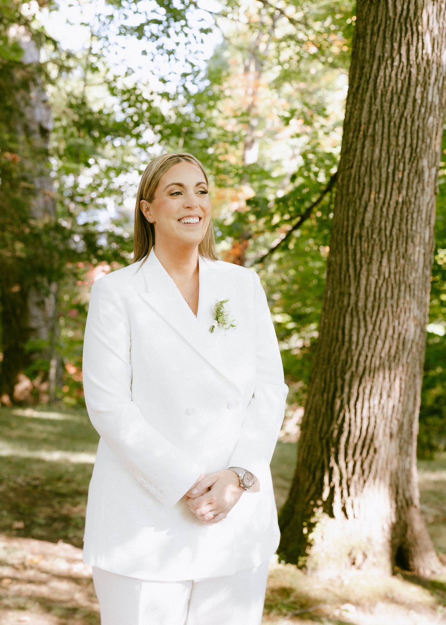 Bride in a white pantsuit smiling outdoors among trees on her wedding day.