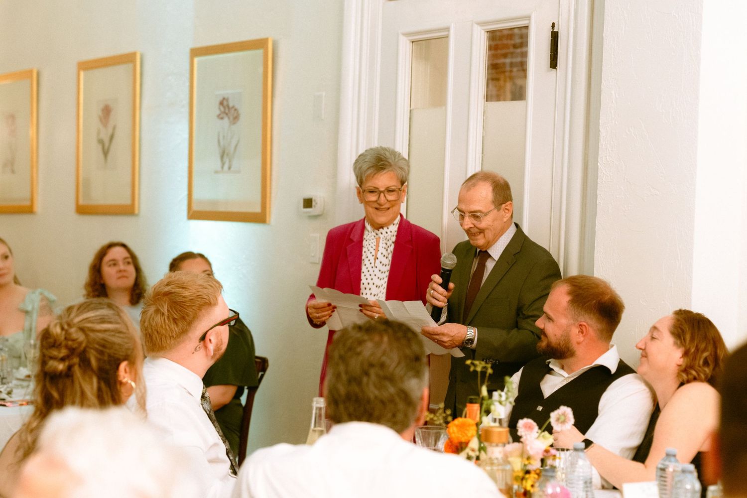 Couple reading a heartfelt speech to laughing wedding reception guests at dinner.