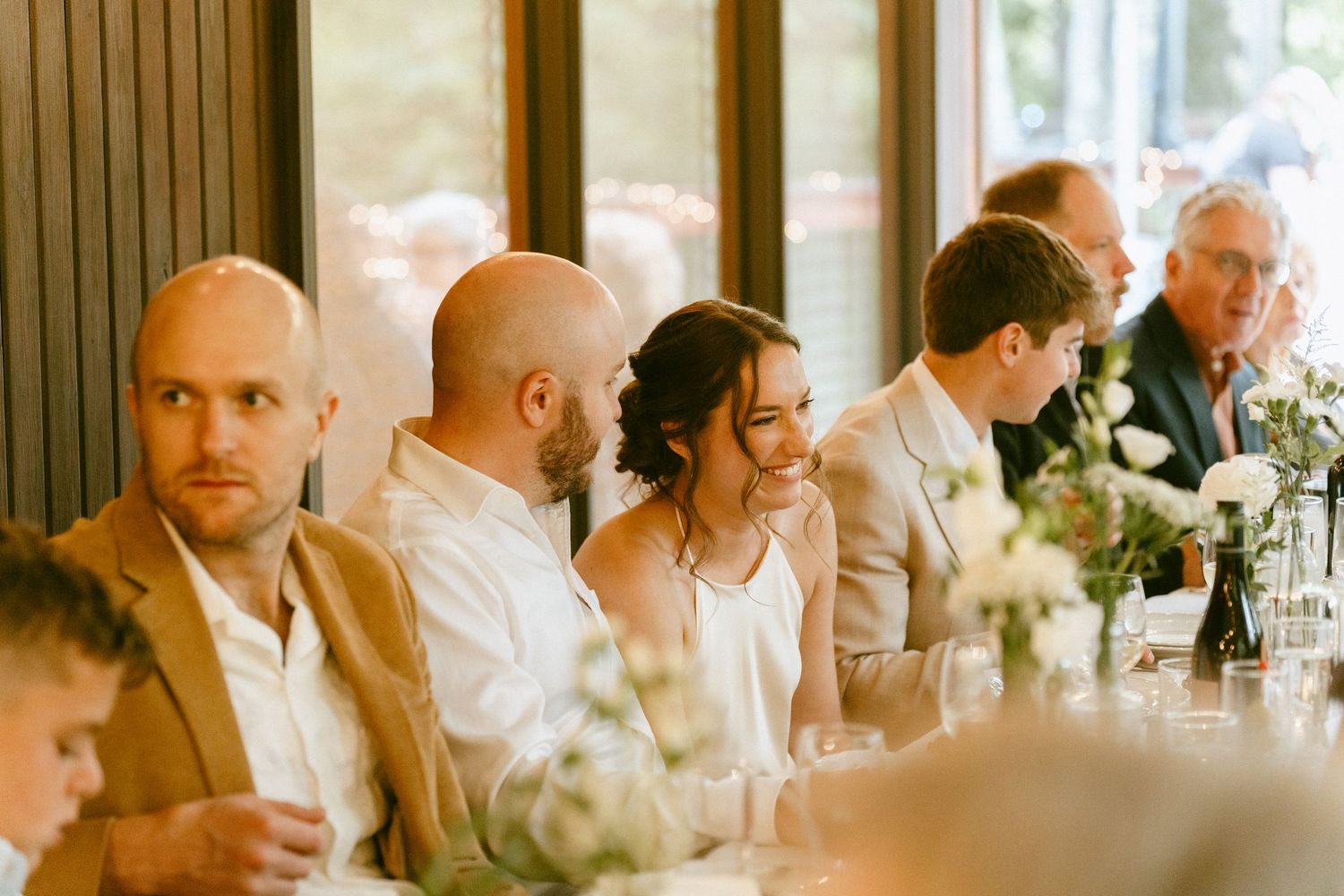 Wedding guests laughing and smiling at a long reception dinner table with white floral centerpieces.