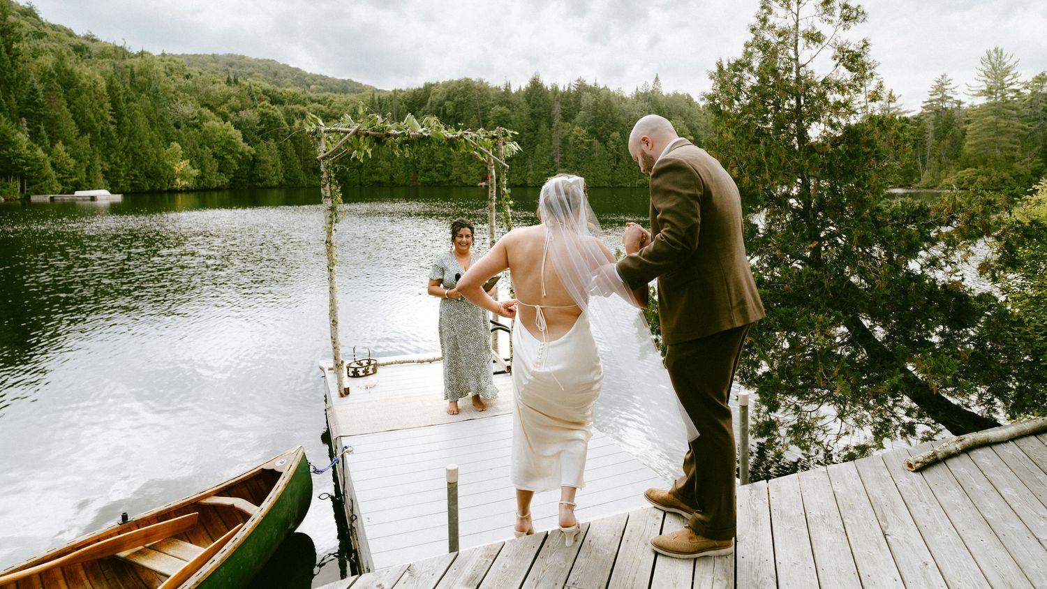Bride walks down the aisle on a scenic lakeside dock with a wooden canoe nearby.