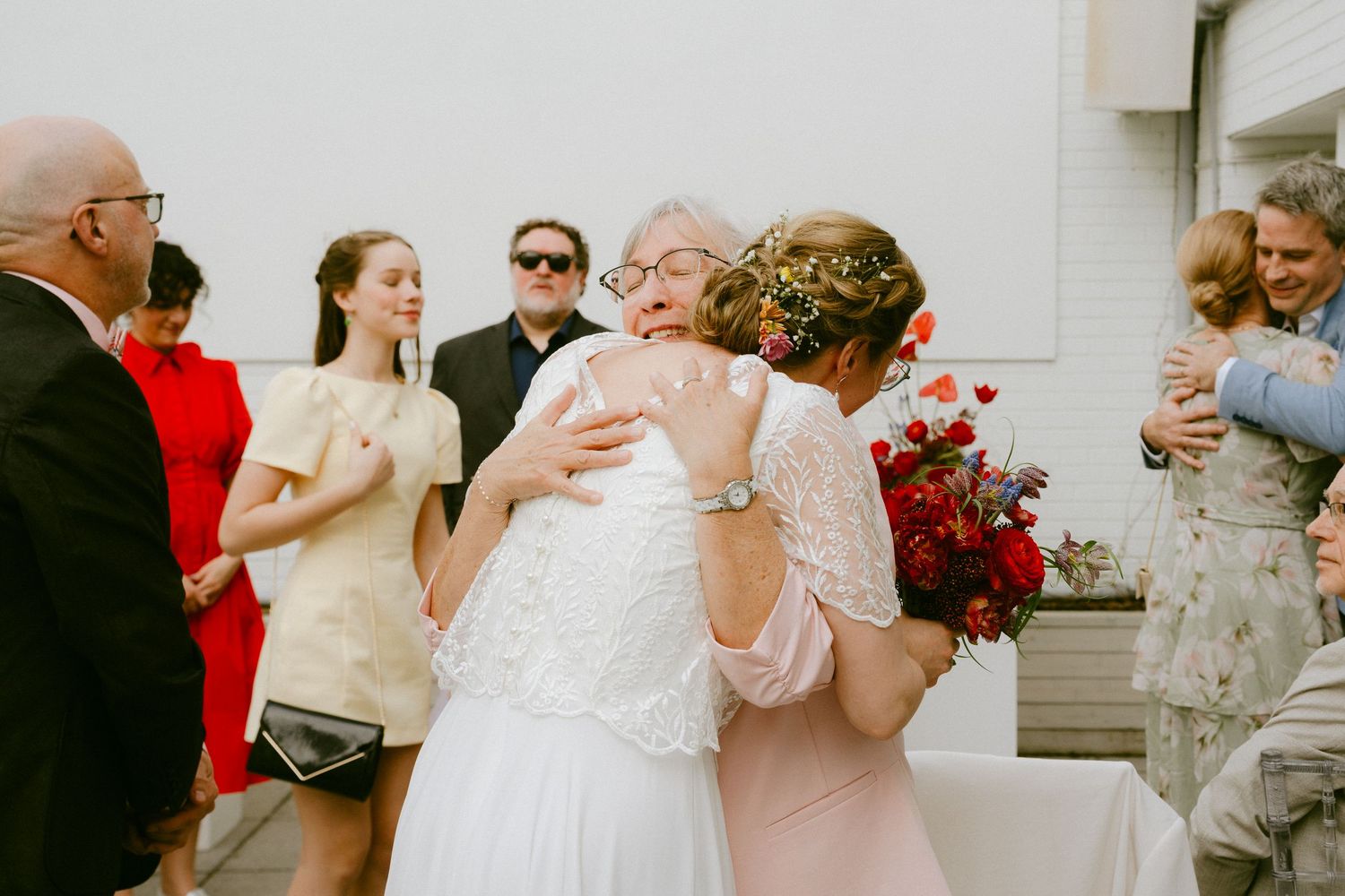 Bride in lace wedding dress embracing guest while holding red bouquet after outdoor wedding ceremony.
