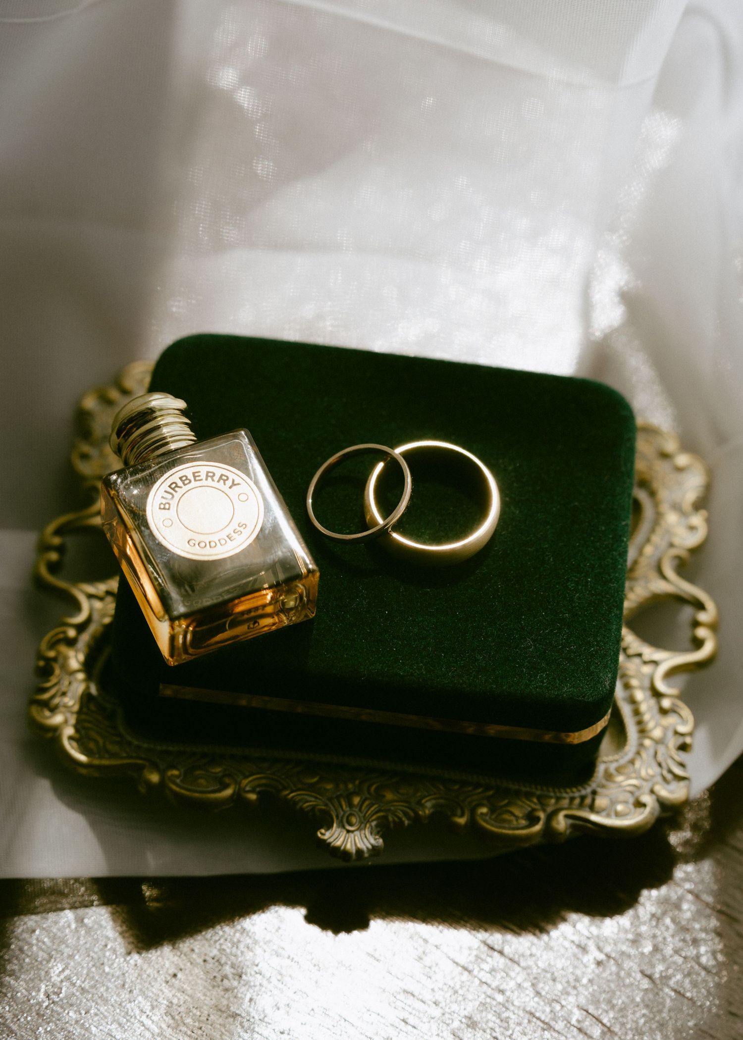 Gold wedding bands and Burberry perfume bottle arranged on a green velvet tray.