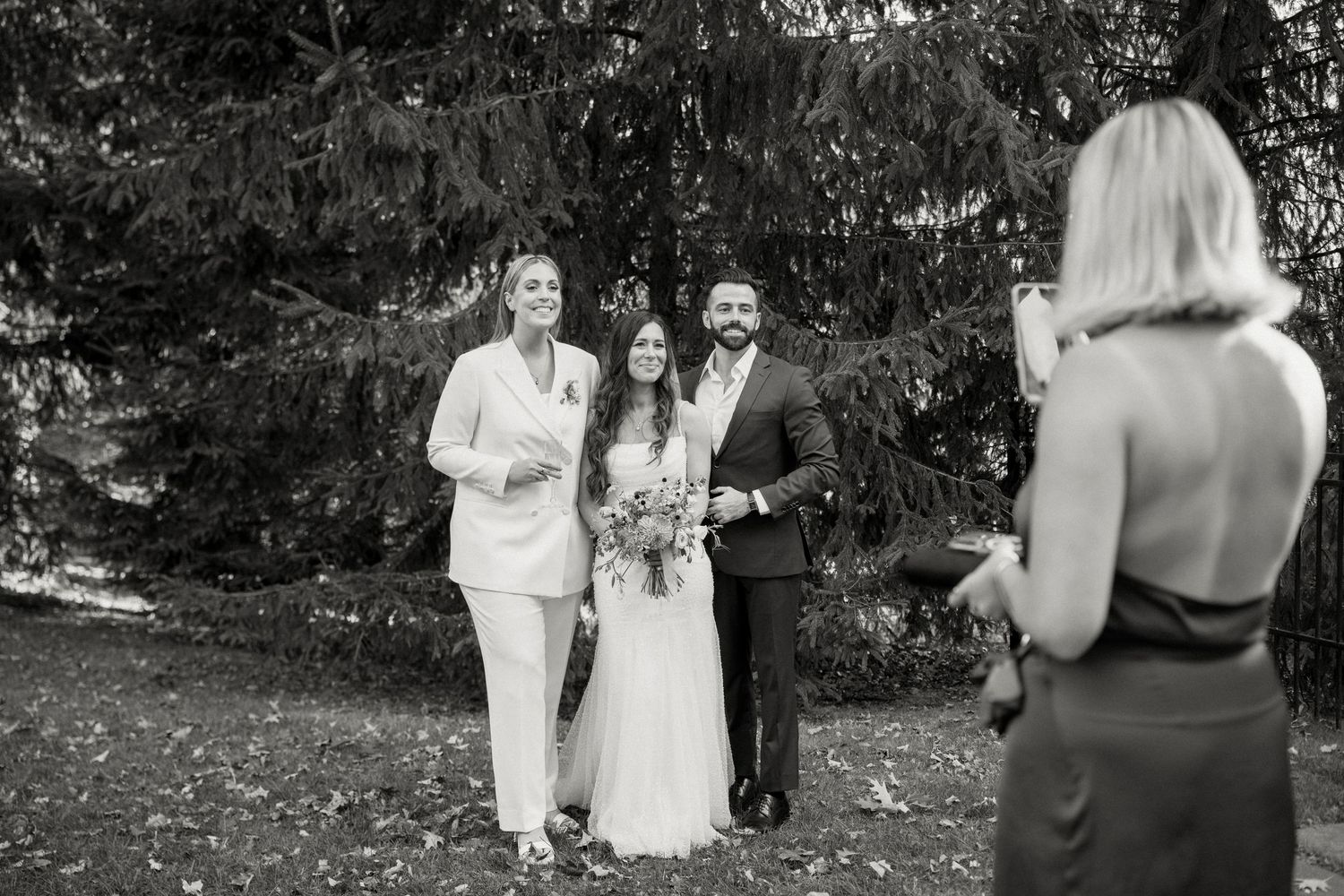 Black and white photo of wedding couple posing with officiant while guest takes their photo.