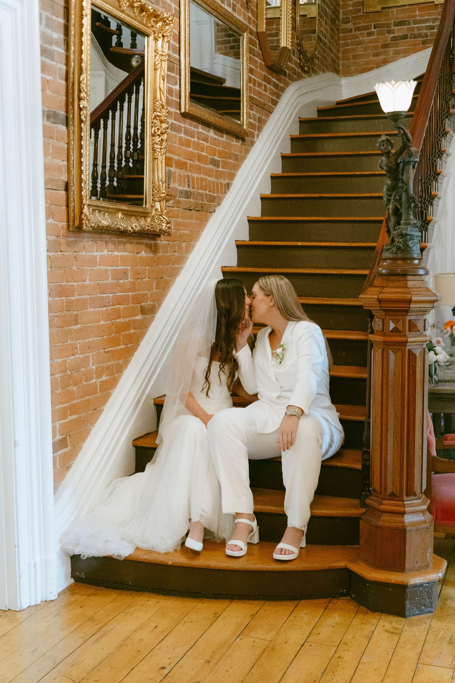 Two brides kiss while seated on wooden staircase with brick wall and gold mirrors.