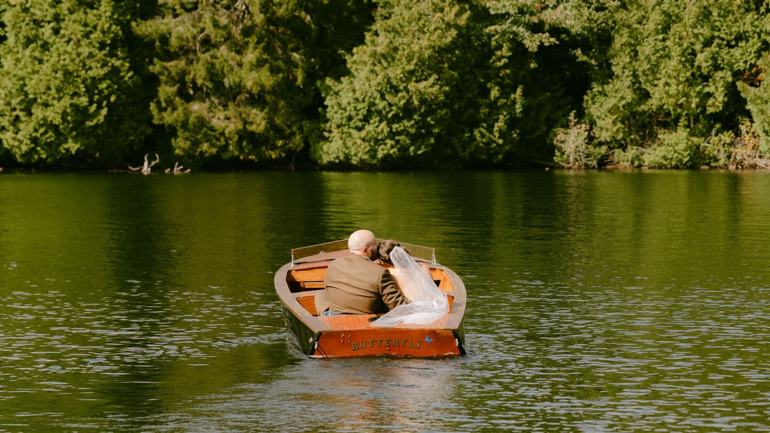 Bride and groom share a romantic moment in a wooden rowboat on a calm, tree-lined lake.