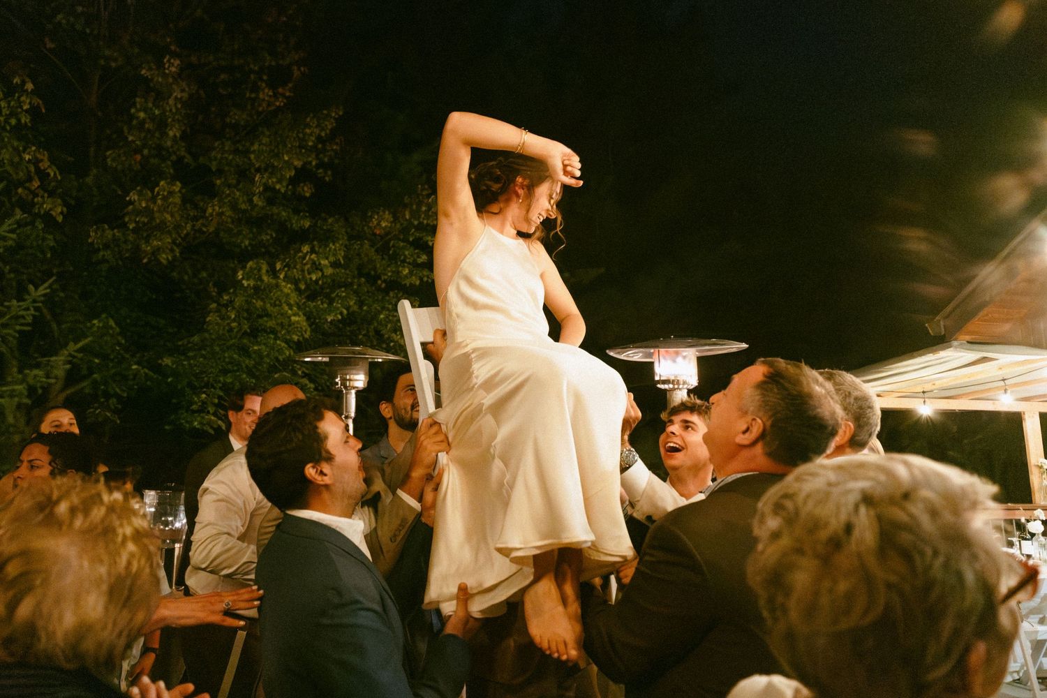 Bride lifted by wedding guests during reception hora dance outdoors at night.