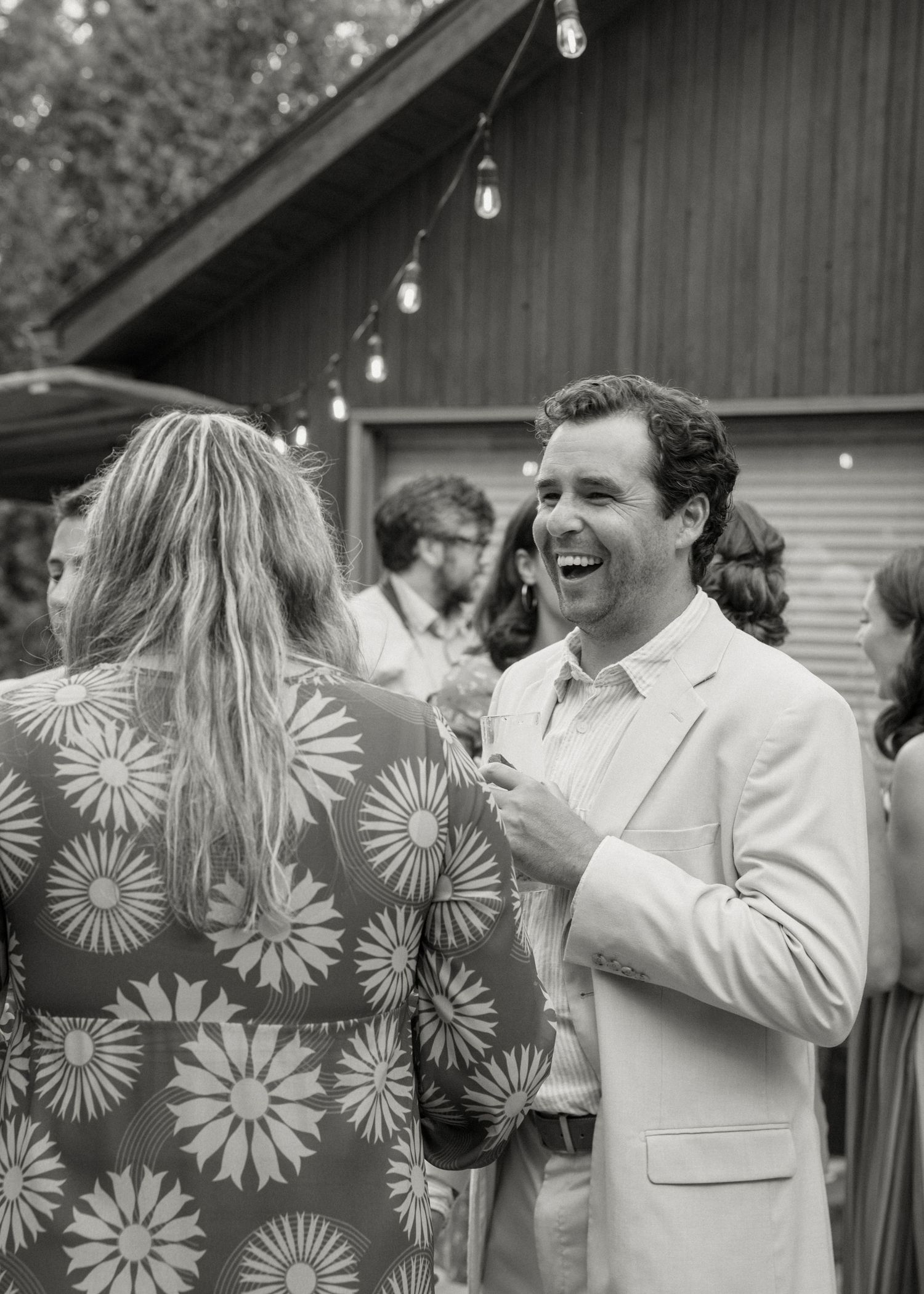 Black and white photo of groom in white suit laughing with female guest at outdoor reception.