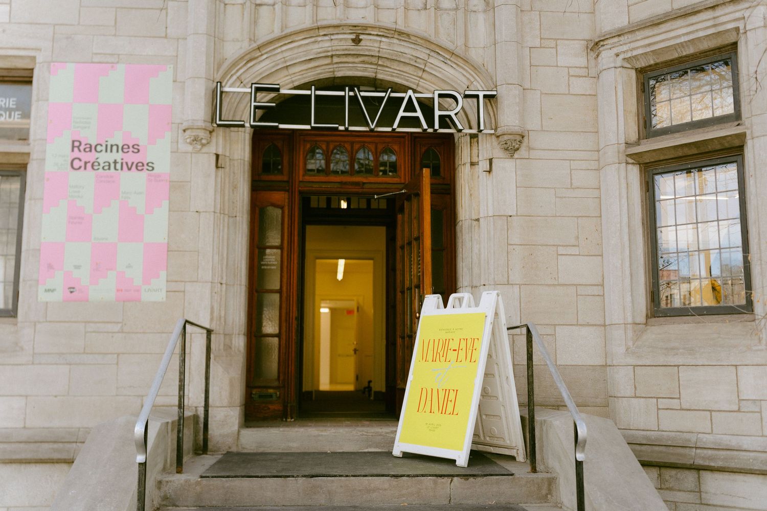 Exterior of Le Tivart art gallery with stone facade, wooden doors, and colorful signage including Racines Creatives poster.