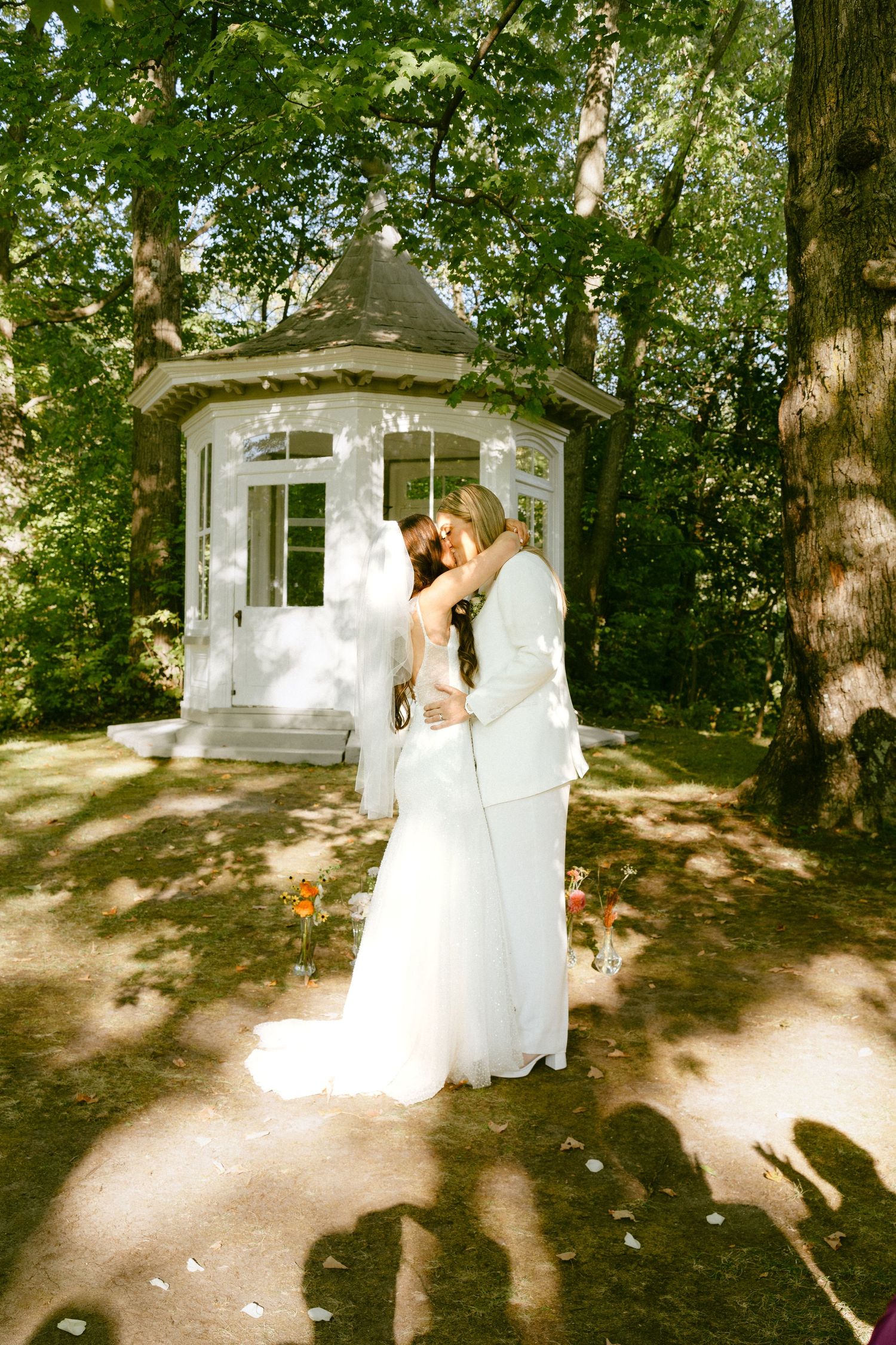 Two brides share a kiss in front of a white gazebo surrounded by lush green trees.