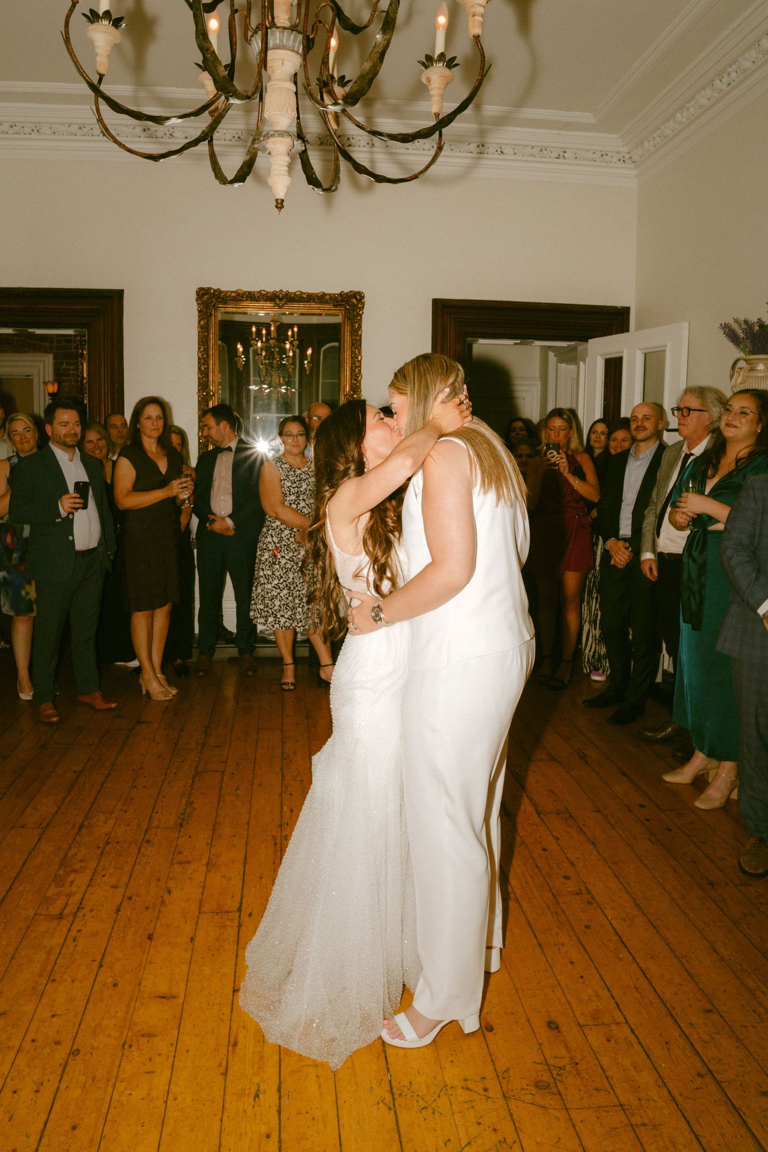 Two brides share a romantic first dance kiss on hardwood floor beneath chandelier at wedding reception.