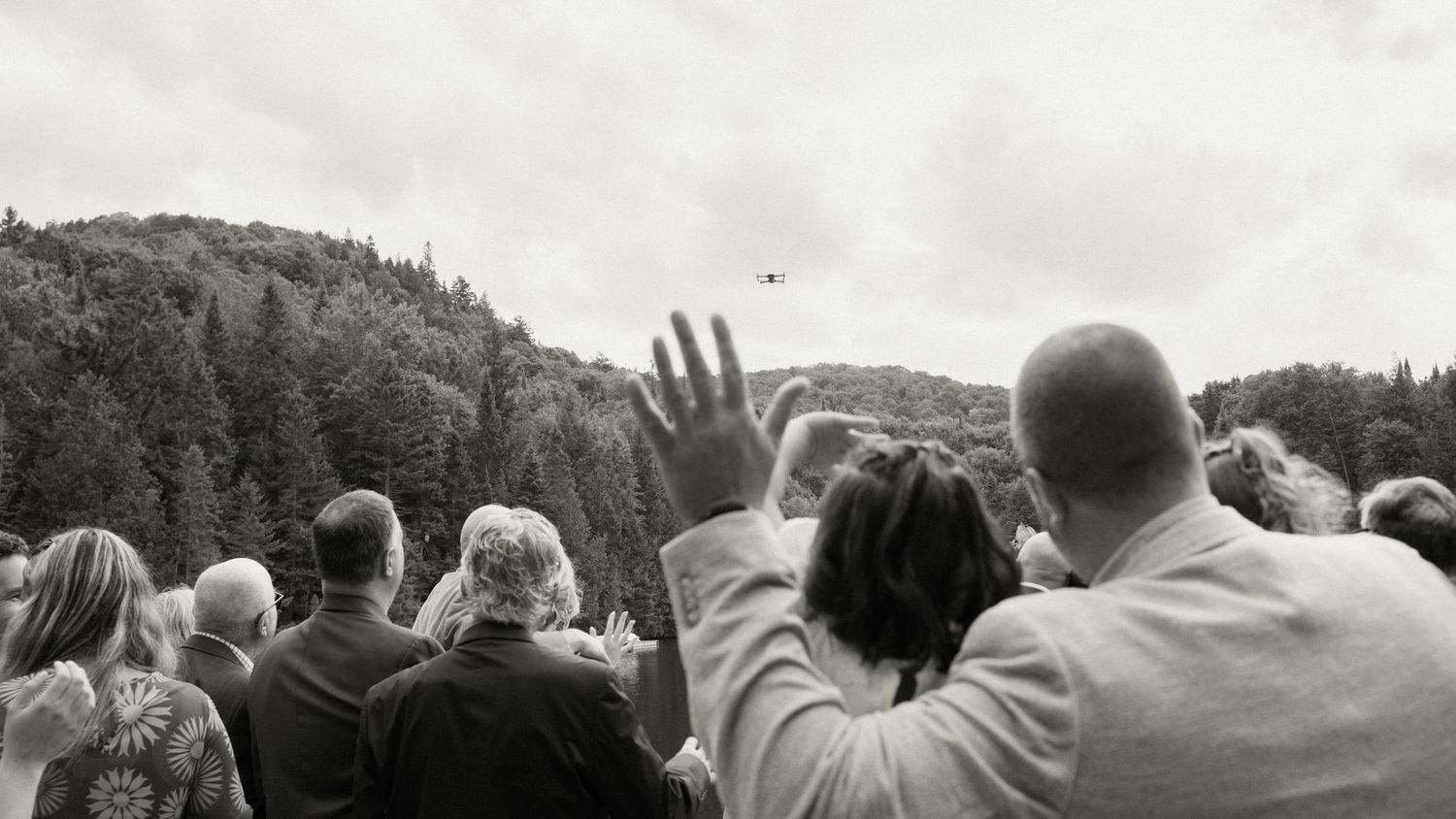 Crowd of wedding guests waving at a drone flying over forested hills in a black and white outdoor photo.