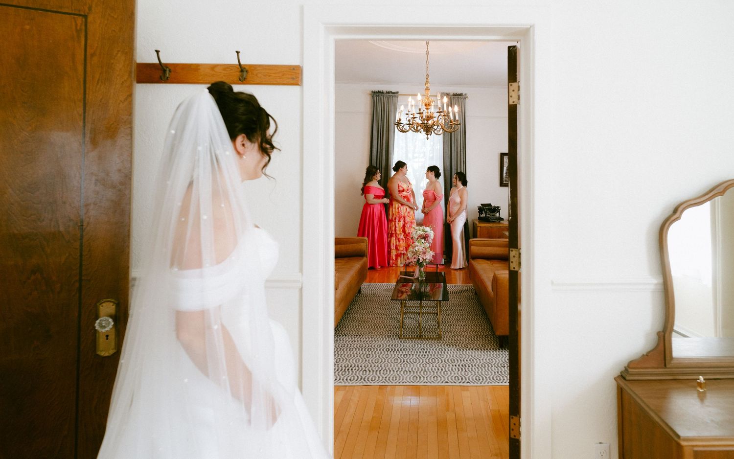 Bride in white gown and veil stands near door while bridesmaids in red dresses gather in background.