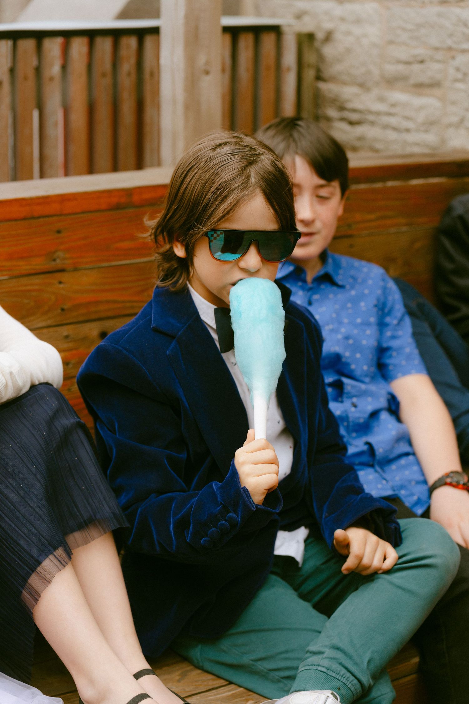 Stylish child in sunglasses and blue velvet blazer eating blue cotton candy at wedding.
