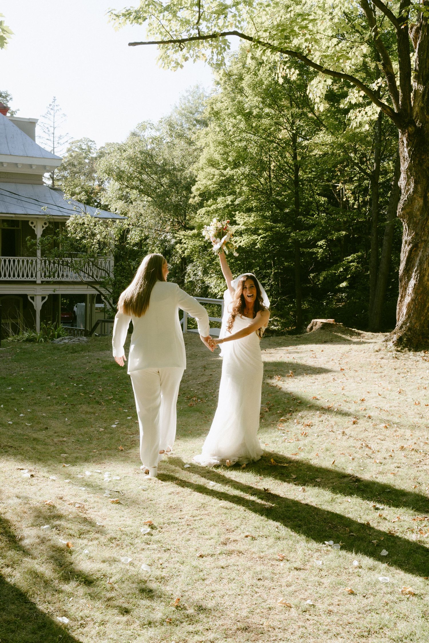 Two brides in white dance playfully on a sunny lawn with a Victorian house in the background.