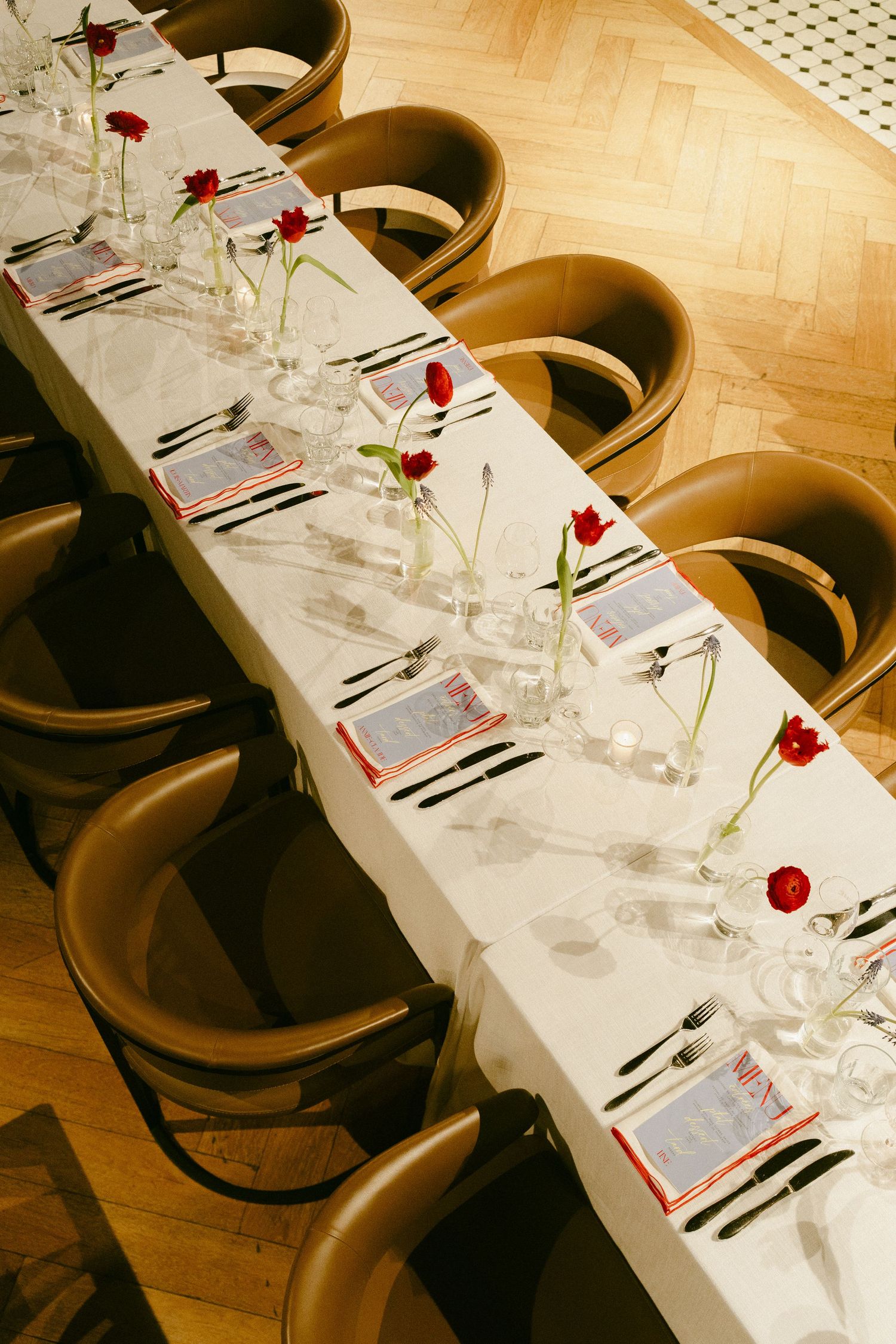 Elegant long dining table set with white linen, red tulips, crystal glasses, and black cutlery for a formal event.