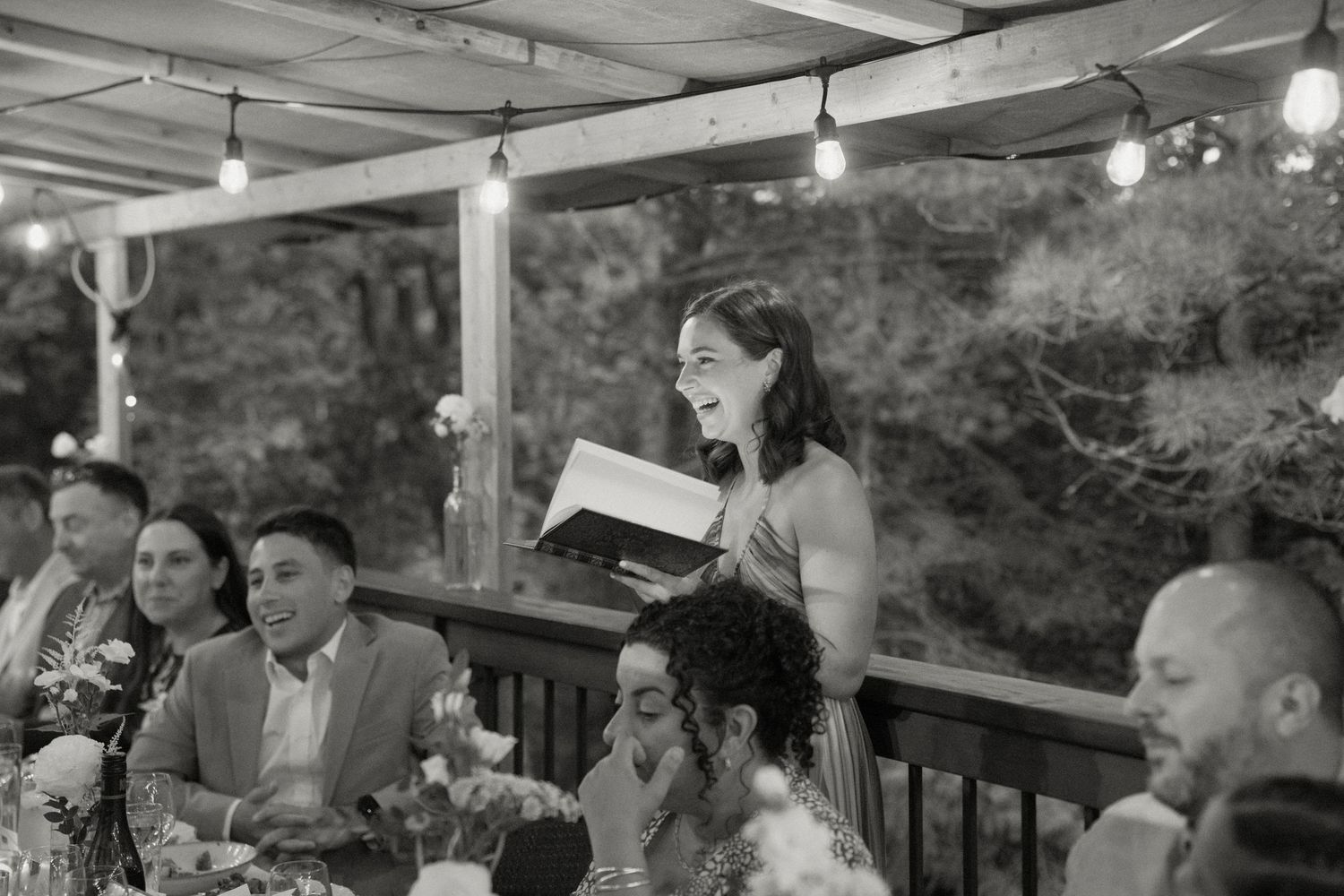 Woman laughing while giving a speech at an outdoor wedding reception under string lights.