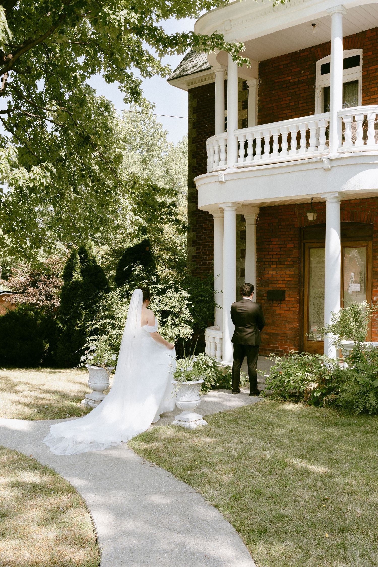 Bride in white gown with cathedral veil approaches groom standing at door of brick manor house.