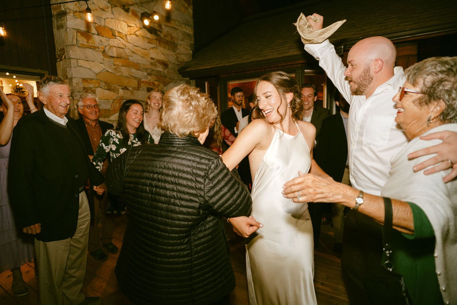 Bride in white gown dancing joyfully with elderly guests at wedding reception.