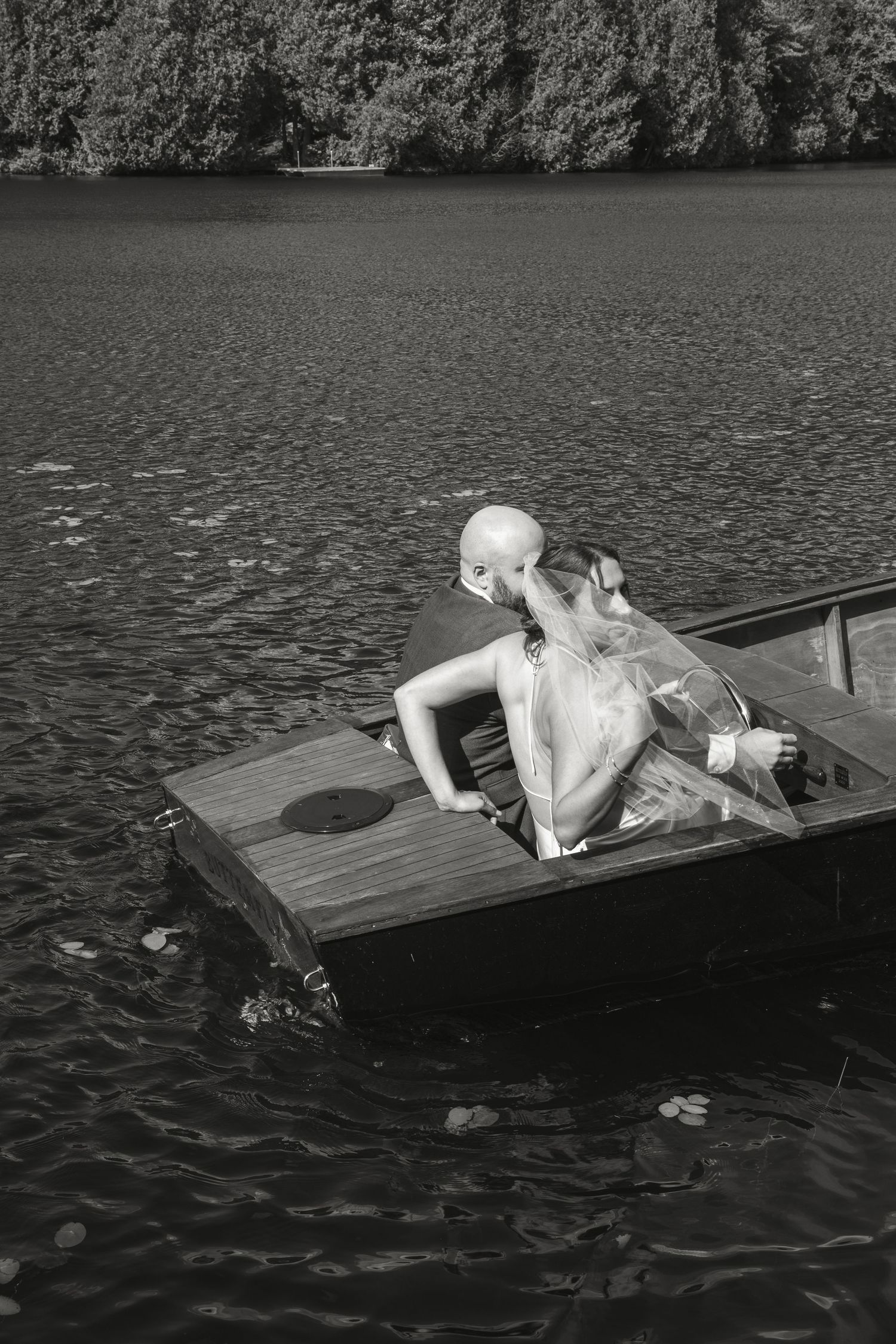Black and white photo of bride and bald groom sitting together in a wooden boat on a lake.