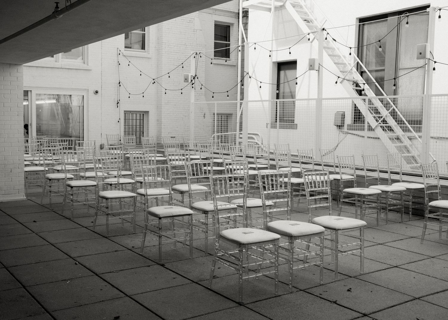 Black and white photo of empty outdoor courtyard with rows of chairs and string lights for an event setup.