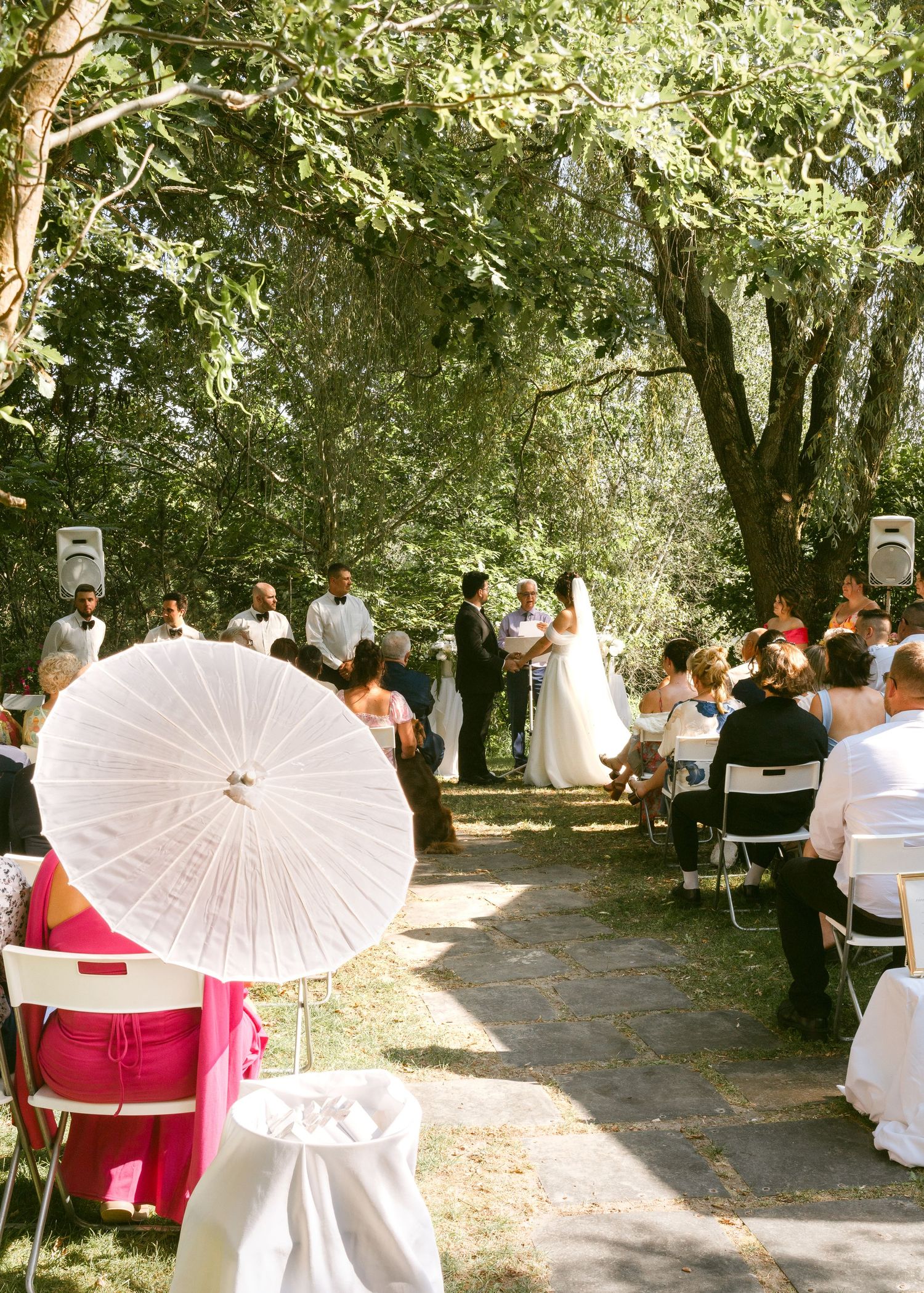 Guests seated at outdoor wedding ceremony under trees with white parasol in foreground.