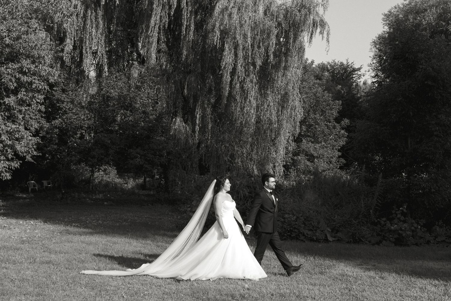 Black and white photo of bride and groom walking hand in hand near a weeping willow tree.