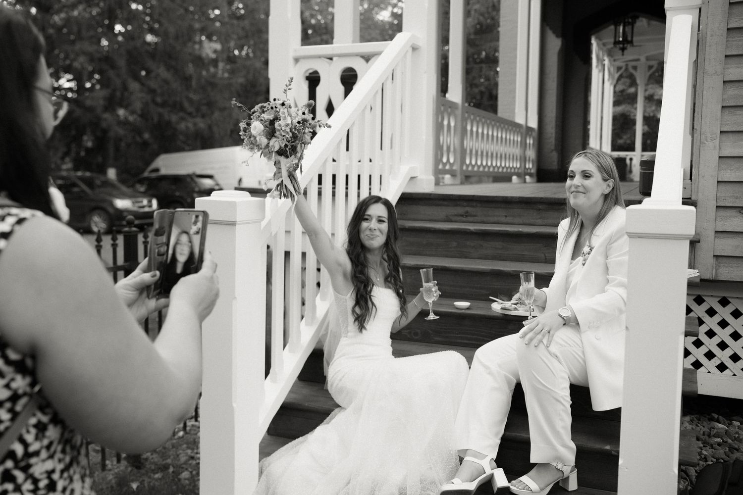 Bride in white dress and partner in white suit sit on porch steps holding champagne while guest photographs them.