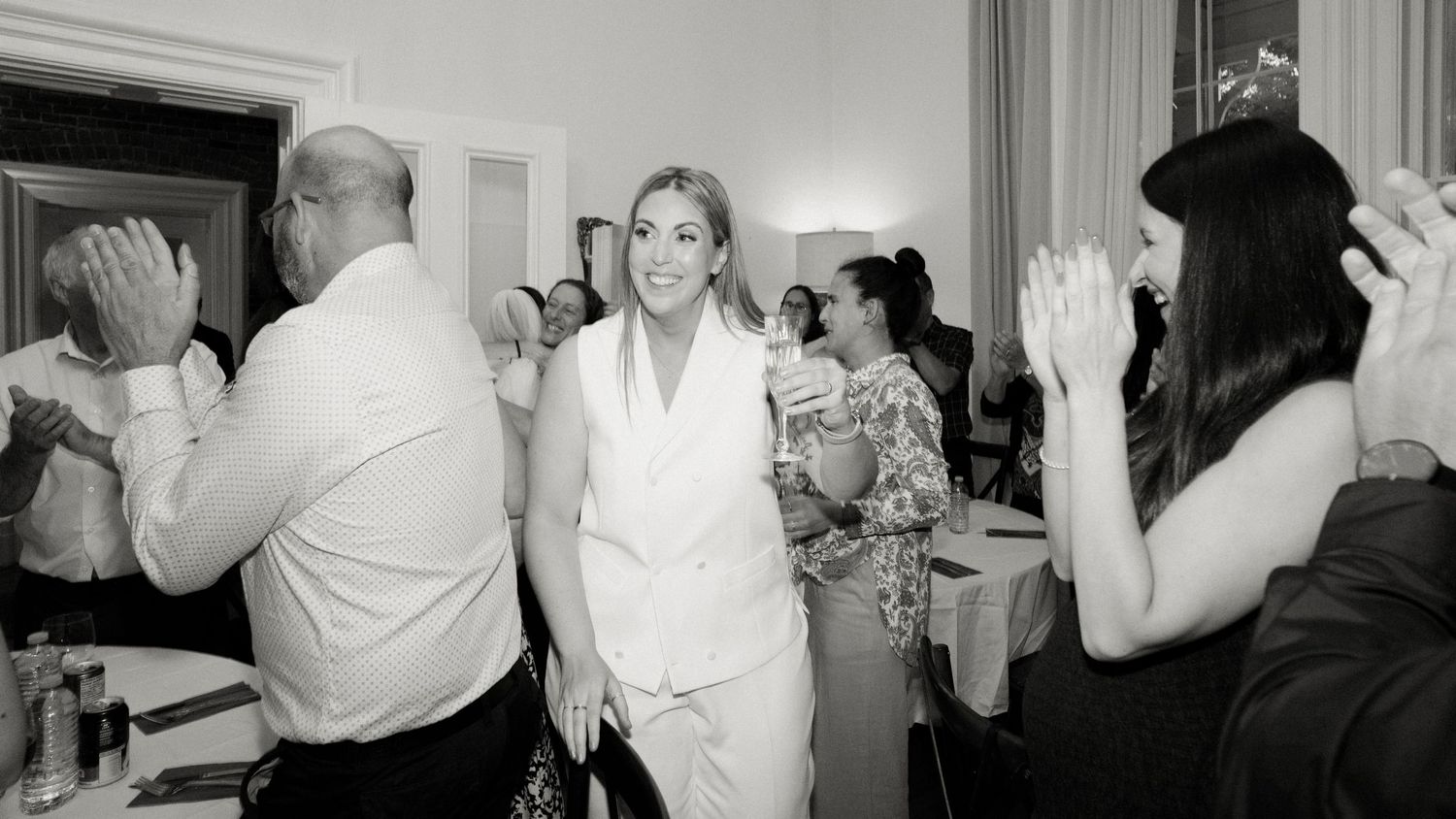 Woman in white suit walking through applauding wedding guests in elegant reception hall.