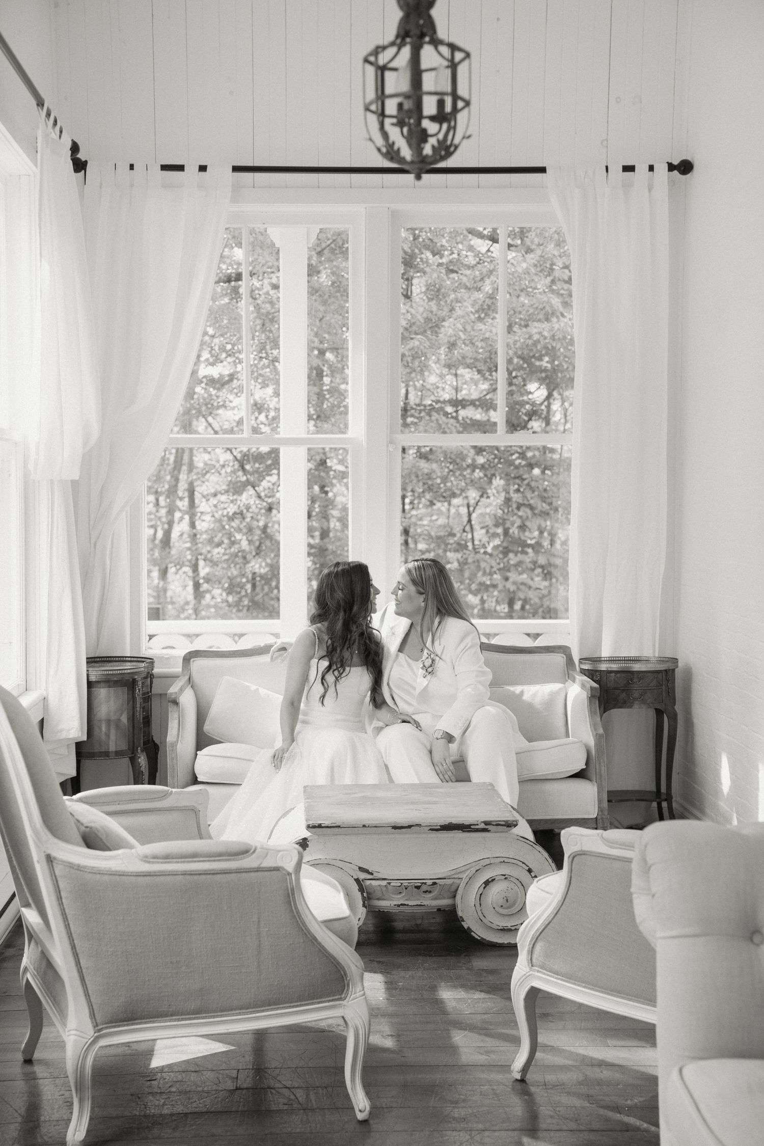 Two brides sit together on sofa in elegant white room with large windows.