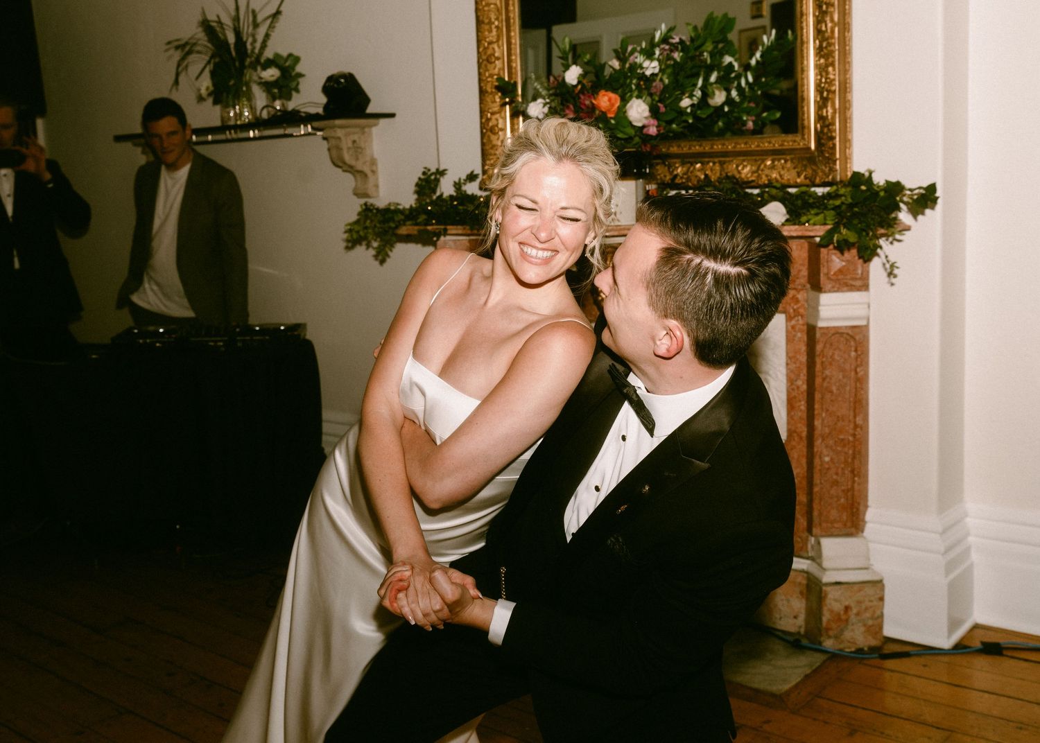 Bride in white satin gown laughing with groom in tuxedo during a playful first dance at their reception.