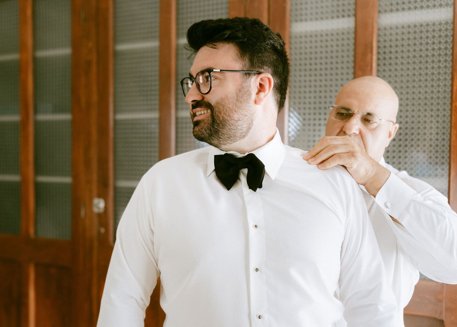 A bearded groom in a white shirt and black bow tie gets ready for his wedding.