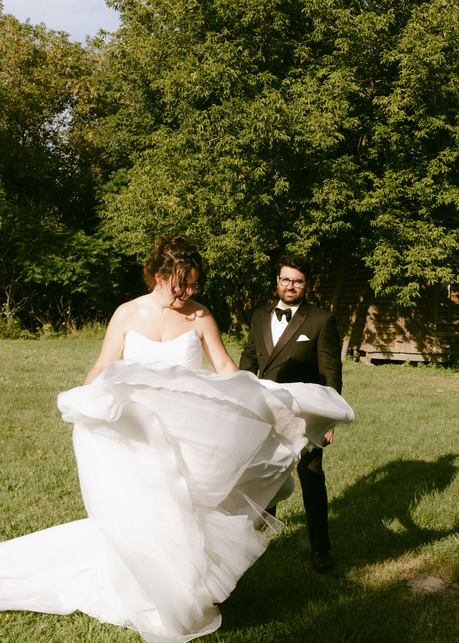 Bride twirls her white wedding gown while groom watches in a sunny green outdoor setting.