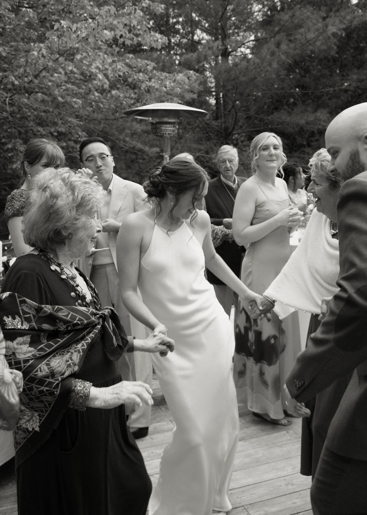 Black and white photo of bride in white gown dancing with elderly woman at outdoor wedding reception.